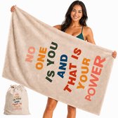 Een lachende vrouw houdt een beige handdoek omhoog met de tekst 'NO ONE IS YOU AND THAT IS YOUR POWER' in veelkleurige letters. Een bijpassend tasje ligt naast haar.