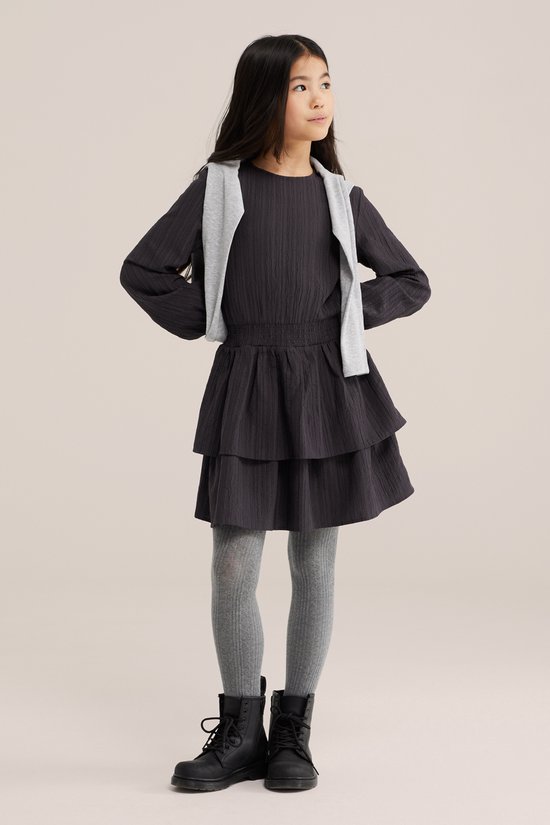 Jong meisje met lang zwart haar draagt een donkergrijze jurk met smocktaille, gelaagde rok, grijze panty en laarzen, met een grijs vestje over de schouders.