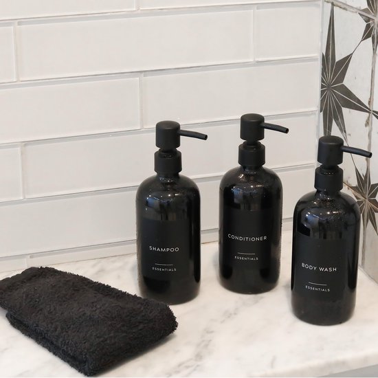 Drie natte, zwarte pompdispensers met labels: BODY WASH, SHAMPOO, CONDITIONER op een marmeren blad.