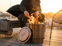 Man voegt hout toe aan brandende Closer Blaze vuurkorf op een houten terras bij zonsondergang. Naast de vuurkorf ligt een gouden deksel.