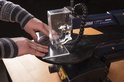 Handen van een persoon die een stuk doorzichtig materiaal op een blauwe FERM figuurzaagmachine plaatst.