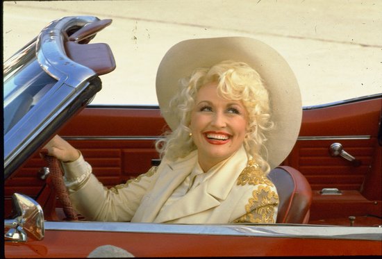 Dolly Parton met blonde krullen, witte hoed en beige jas met gouden details, in een rode cabriolet.