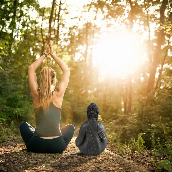 Donkergrijs Boeddhabeeld zittend in een yogahouding naast een vrouw in een bosrijke omgeving.