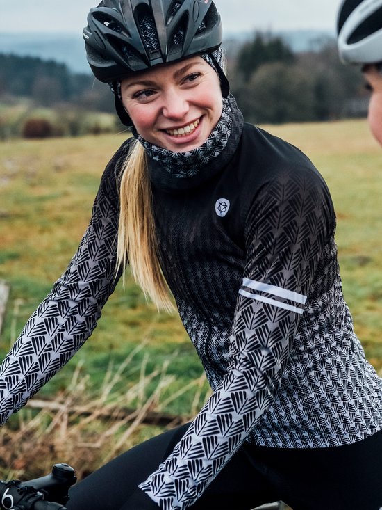 Lachende vrouw op een fiets in een zwart fietsshirt met lange mouwen, een geometrisch grijs-wit patroon en een bijpassende nekwarmer.