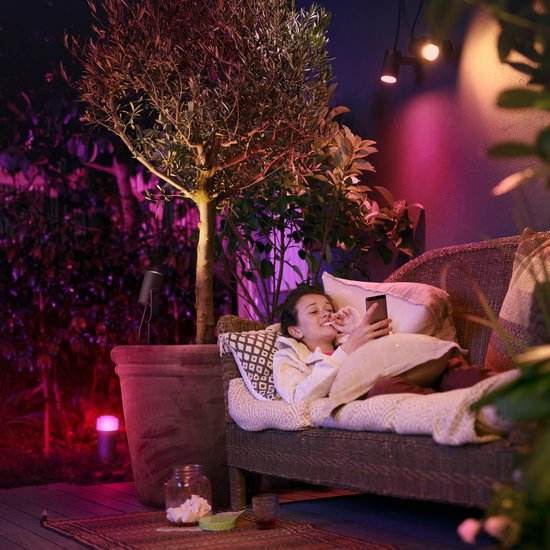 Vrouw ontspant op een loungeset in een tuin die verlicht is met gekleurde Philips Hue spots, met een telefoon in haar hand.