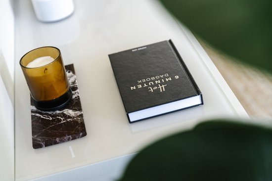 Zwart dagboek met gouden letters "Het 6 minuten dagboek" op een glanzend tafelblad, naast een geurende kaars.