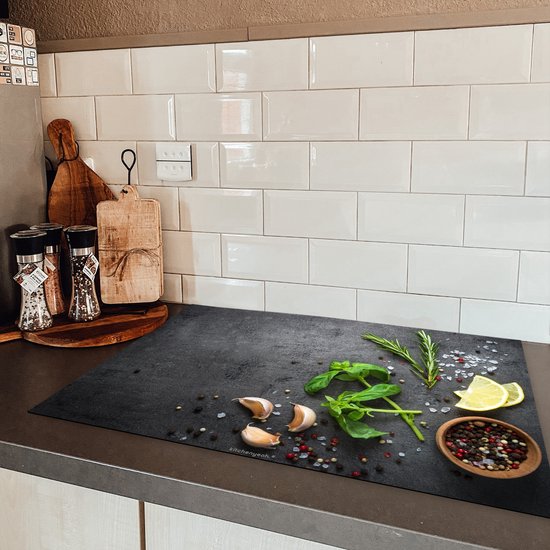 Zwarte inductie beschermer met betonprint, basilicum, rozemarijn, citroenpartjes, knoflook en peperkorrels, in een keuken met witte tegels.
