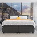 Opgebedde grijze boxspring met witte lakens, oranje plaid, gele kussen en uitzicht op een kuststad.