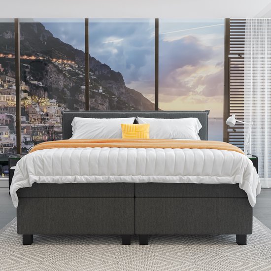 Opgebedde grijze boxspring met witte lakens, oranje plaid, gele kussen en uitzicht op een kuststad.