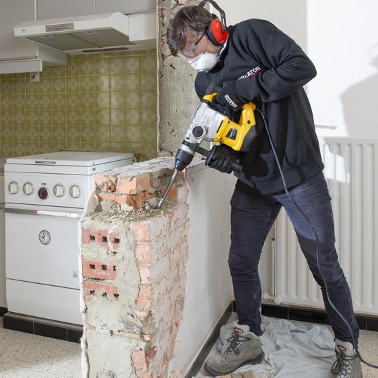 Man met veiligheidsuitrusting die een gele en grijze Powerplus klopboormachine gebruikt om muur af te breken.