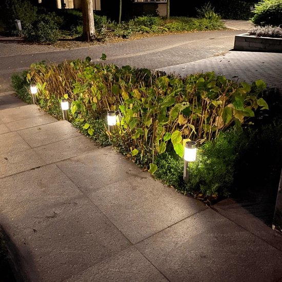Zilverkleurige solar paaltjes met witte verlichting, langs een betegeld pad en struiken in de avond.