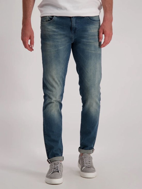 Man draagt een blauwe spijkerbroek met lichte wassing en omgerolde broekspijpen. Slim fit.