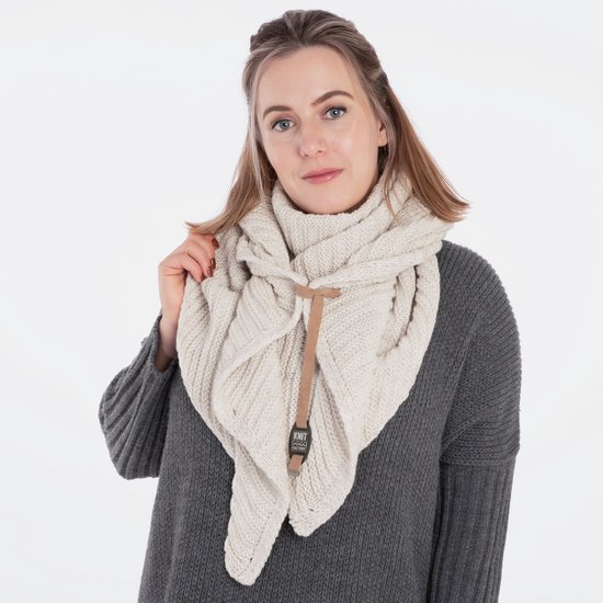 Model draagt een beige gebreide driehoekssjaal over een grijze trui. De sjaal heeft een bruine siersluiting met Knit Factory logo.