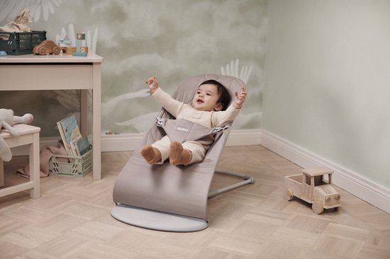 Lachende baby in zandgrijs wipstoeltje met handen omhoog, in een kinderkamer.