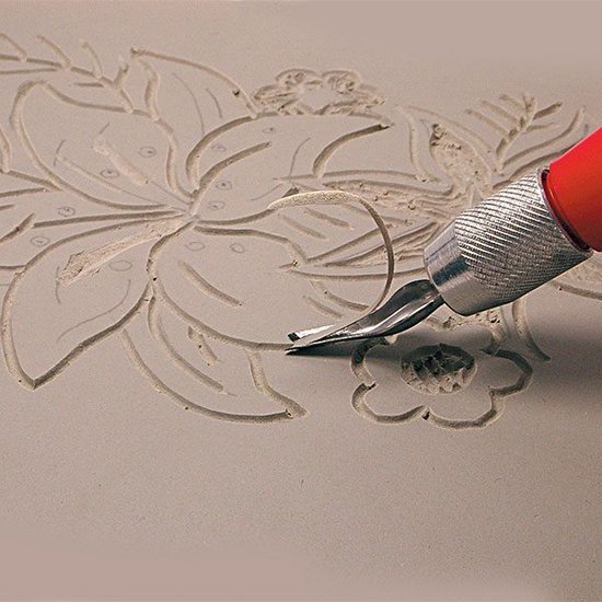 Een hand met een snijgereedschap dat een bloemmotief in grijs linoleum snijdt.