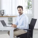Man met een lichtblauwe blouse en beige broek zit op een stoel met een rugkussen, kijkend naar zijn telefoon.
