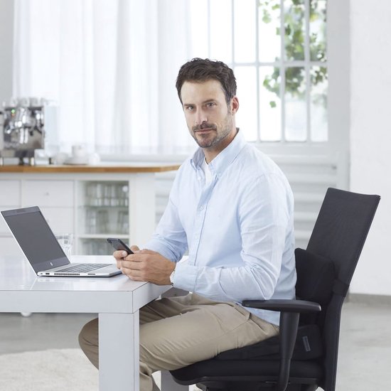 Man met een lichtblauwe blouse en beige broek zit op een stoel met een rugkussen, kijkend naar zijn telefoon.
