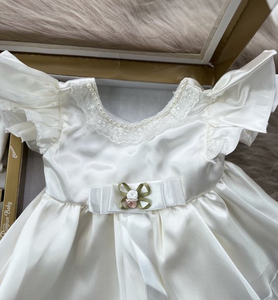 Detail van een babyjurk in ivoorkleur met kanten kraagje en strik met bloemdetail.