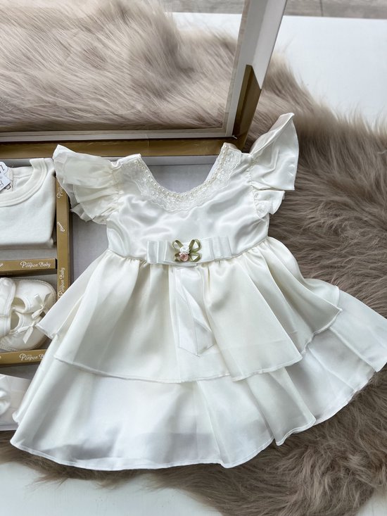 Babyjurk in ivoorkleur met kanten kraagje, strik met bloemdetail en gelaagde rok. Bijpassend rompertje, haarband en schoentjes.