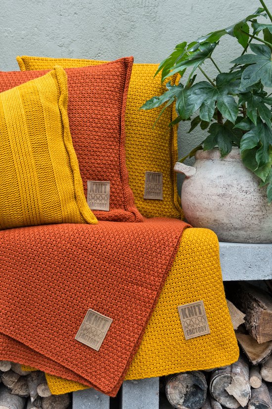 Plaids et coussins tricotés orange et moutarde avec des étiquettes marron, à côté d'une plante verte.