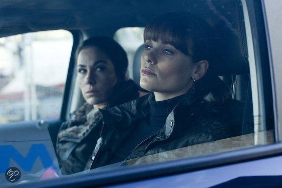 Twee vrouwen in een auto. De voorste vrouw heeft een pony en blauwe ogen, de achterste vrouw heeft donker haar.