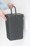 Mains tenant une poubelle Brabantia gris foncé par son anse, près d'un support mural.