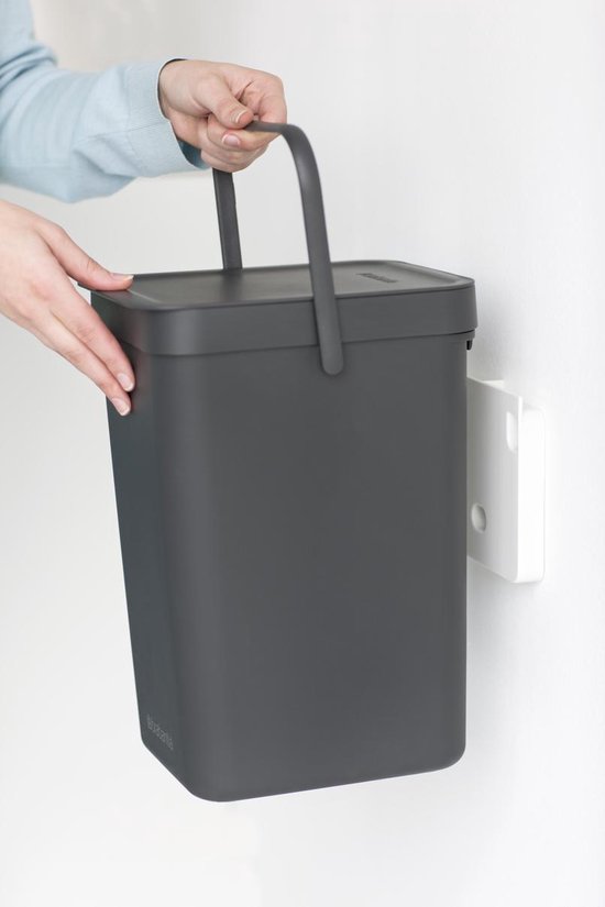 Mains tenant une poubelle Brabantia gris foncé par son anse, près d'un support mural.