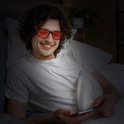 Een lachende man met een witte T-shirt ligt in bed en kijkt op zijn telefoon door een bril met oranje glazen.