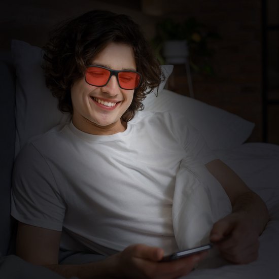 Een lachende man met een witte T-shirt ligt in bed en kijkt op zijn telefoon door een bril met oranje glazen.