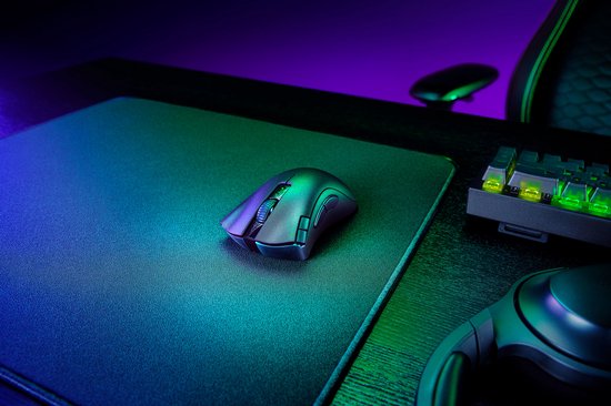 Een donkergrijze gamingmuis op een zwart gamingmuismat, met een verlicht toetsenbord en een gamingstoel op de achtergrond.