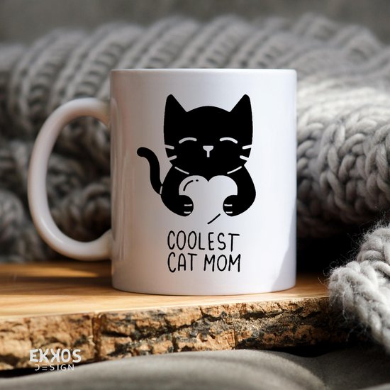 Witte mok met zwarte print van een kat die een hart vasthoudt met de tekst "COOLEST CAT MOM". Achtergrond met dik gebreide stof.