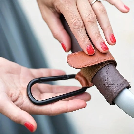 Handen met rode nagellak bevestigen een bruine kunstlederen kinderwagenhaak met zwarte karabijnsluiting.