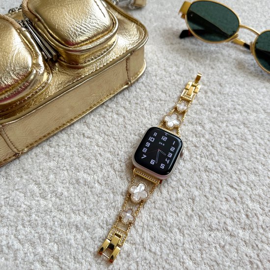 Gouden Apple Watch bandje met klaver en parel motief, naast een gouden handtas en zonnebril.