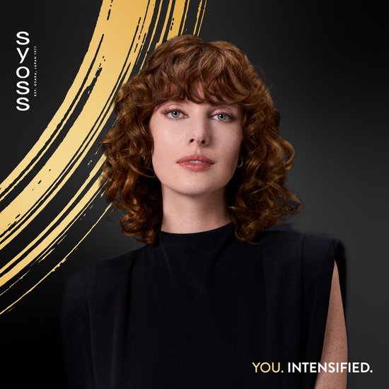 Model met golvend roodbruin haar, gekleed in zwart, poseert voor de camera. Gouden accent op de achtergrond.