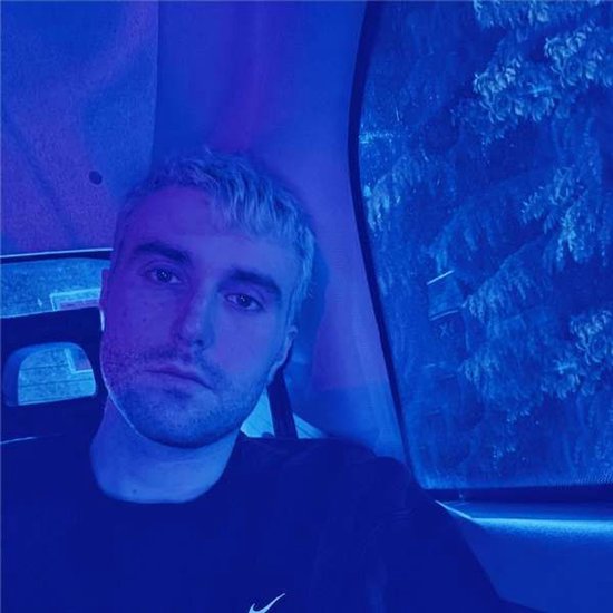 Een man met kort blond haar en grijze baard, deels zichtbaar in een auto, met blauwe verlichting.