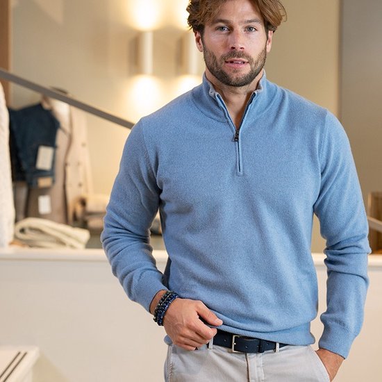 Homme portant un pull bleu clair à demi-fermeture éclair, un pantalon beige et un bracelet bleu.