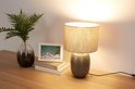 Grijze tafellamp met beige kap en decoratieve voet, op een houten tafel naast een vaas en boeken.