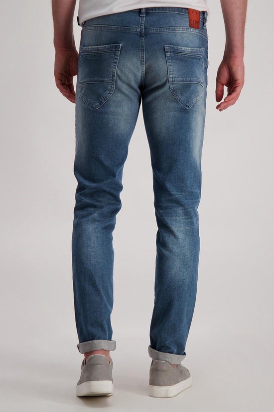 Man van achteren gezien, draagt een blauwe spijkerbroek met opgerolde broekspijpen en grijze sneakers.