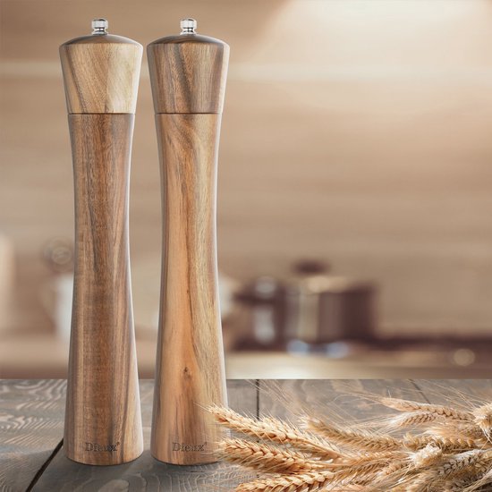 Twee slanke, hoge peper- en zoutmolens van natuurlijk acaciahout op een houten tafel met korenaren op de voorgrond. De achtergrond is onscherp.
