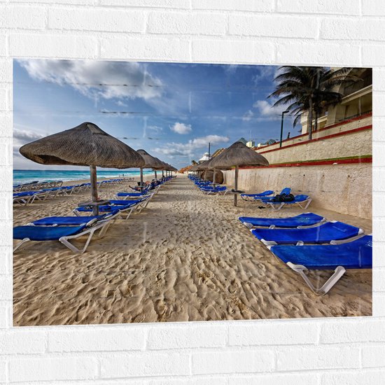 WallClassics - Muursticker - Blauwe Ligbedden op Strand met Rieten Parasols - 100x75 cm Foto op Muursticker