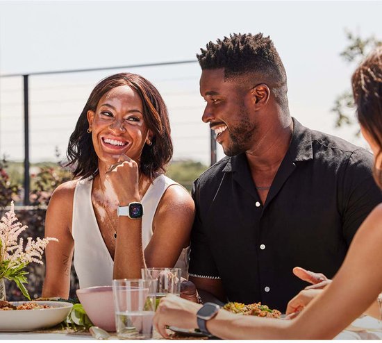 Vrouw met vitiligo lacht en draagt een goudkleurig Fitbit Sense 2 smartwatch met lichtblauw bandje tijdens een maaltijd buiten.
