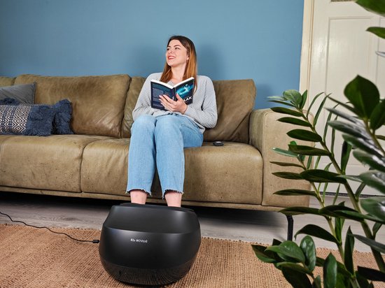Vrouw op een bank leest een boek terwijl haar voeten in een zwart voetmassageapparaat staan.