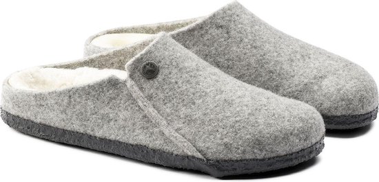 Een paar lichtgrijze vilten Birkenstock pantoffels met schapenvacht voering en zwarte zolen, gezien vanuit een hoek.