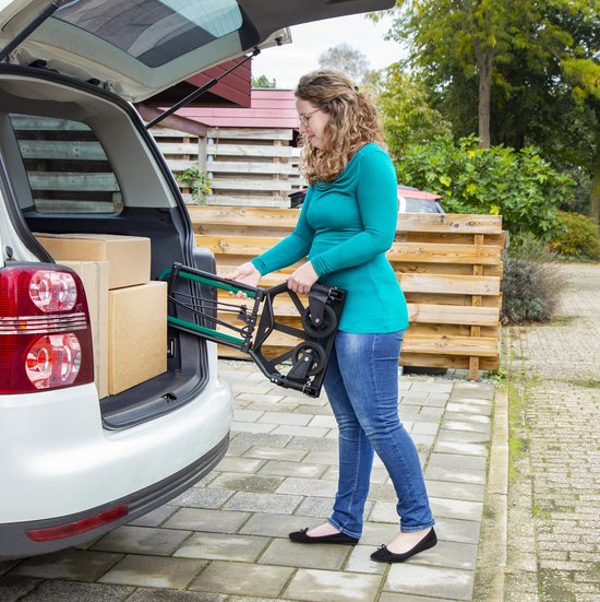 Een vrouw laadt een opvouwbare steekwagen met groene handgrepen in de kofferbak van een auto, naast twee dozen.