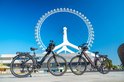 Twee elektrische fietsen, één zwart en één paars, voor een reuzenrad op een zonnige dag.