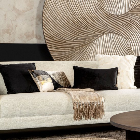 Een beige bank met diverse kussens in zwart, wit en met patronen, een beige plaid met franjes en een grote decoratieve wandsculptuur.