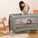 Een vrouw kijkt lachend naar een slapende baby in een lichtgroen campingbedje met gaas zijkanten.