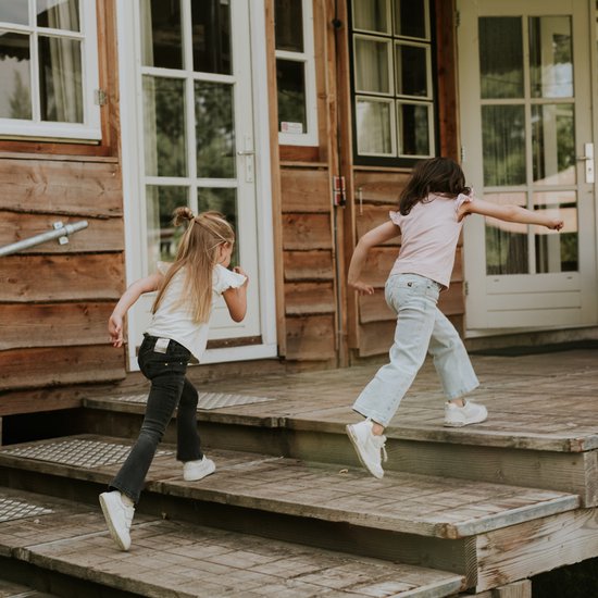 Twee meisjes rennen op een houten trap, één in een donkergrijze flared jeans en de ander in een lichte jeans.