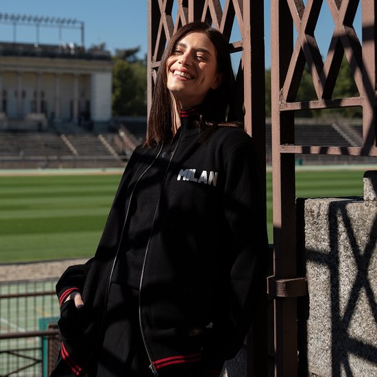 Vrouw draagt een zwarte retro voetbalvest met 'MILAN' opdruk, staand buiten bij een stadion.
