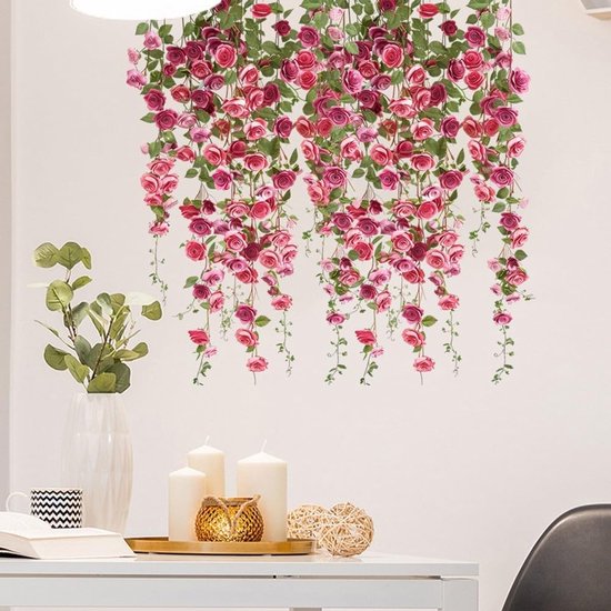Hangende roze en rode rozen met groene bladeren op een witte muur, boven een tafel met decoraties.